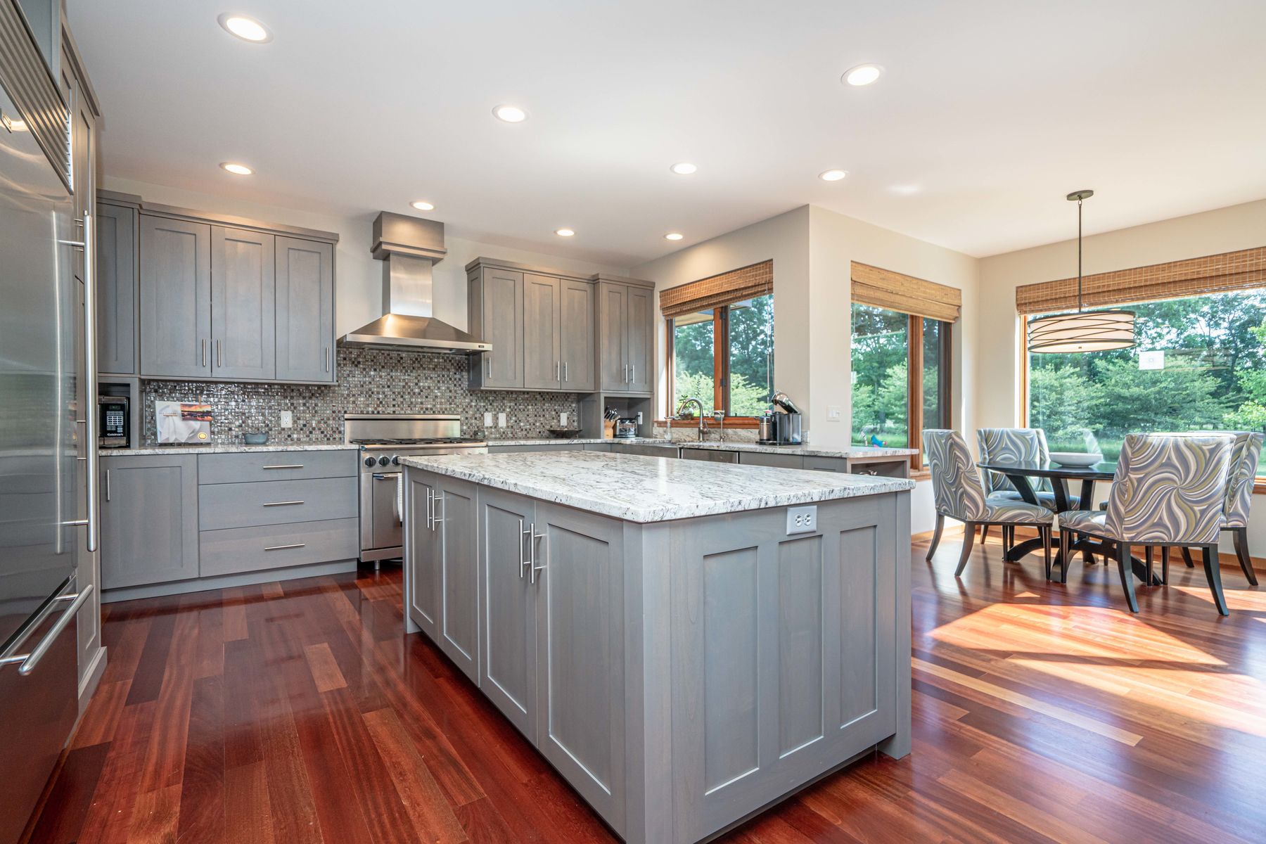 A kitchen with gray cabinets, a stone-topped island and wood floors, opening to a sunny dining area